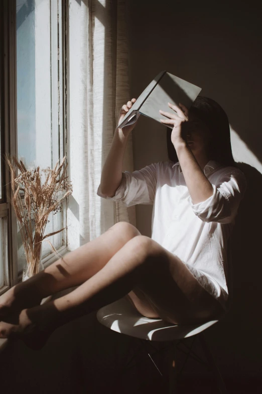 a woman sitting on a chair reading a book, a picture, unsplash contest winner, aestheticism, halo over her head, window light, wearing a light shirt, half body photo