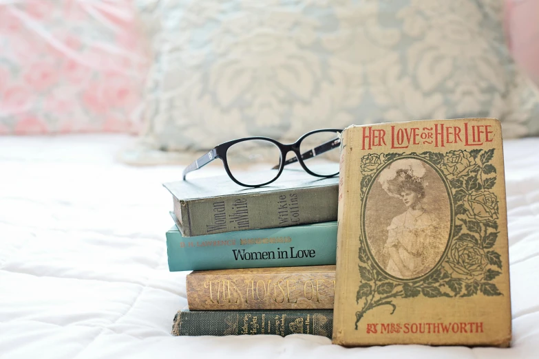 a stack of books sitting on top of a bed, inspired by Cassandra Austen, romanticism, blue rimmed glasses, vintage vibe, style of lady frieda harris, romantic lead