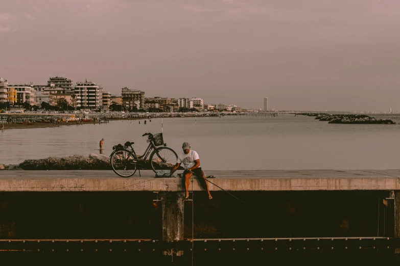 a man sitting on a dock next to a bike, pexels contest winner, happening, the sea seen behind the city, complex and desaturated, brown, humid evening