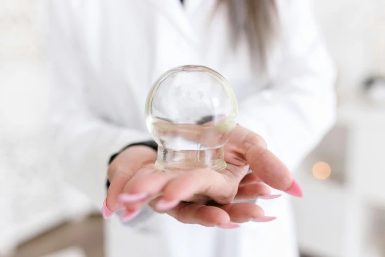 a close up of a person holding a glass object, wearing lab coat and a blouse, the orb of dreams, clear liquid, professional product shot