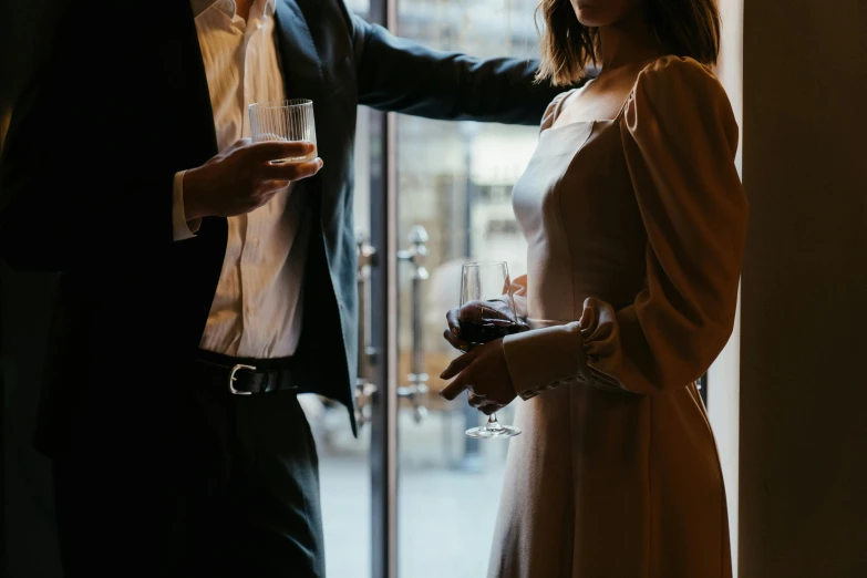 a man standing next to a woman holding a glass of wine, by Lee Loughridge, trending on pexels, renaissance, subject detail: wearing a suit, lit from behind, romantic lead, looking outside