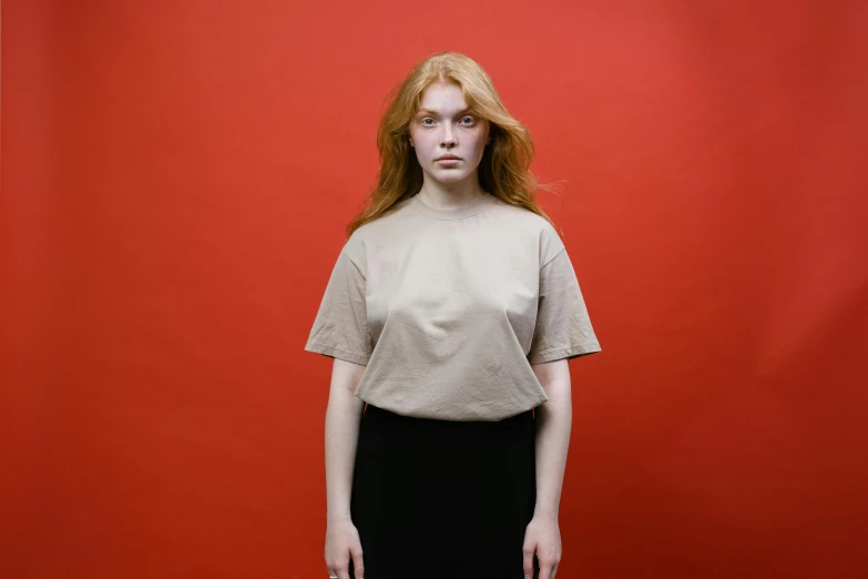 a woman standing in front of a red wall, a character portrait, inspired by Elsa Bleda, pexels contest winner, renaissance, wear's beige shirt, on grey background, sadie sink, red t-shirt