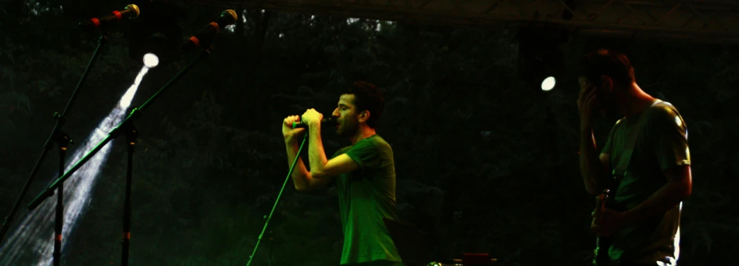 a couple of men standing next to each other on a stage, an album cover, by Attila Meszlenyi, pexels, in front of a forest background, greenish lighting, in an action pose, grainy low quality photograph