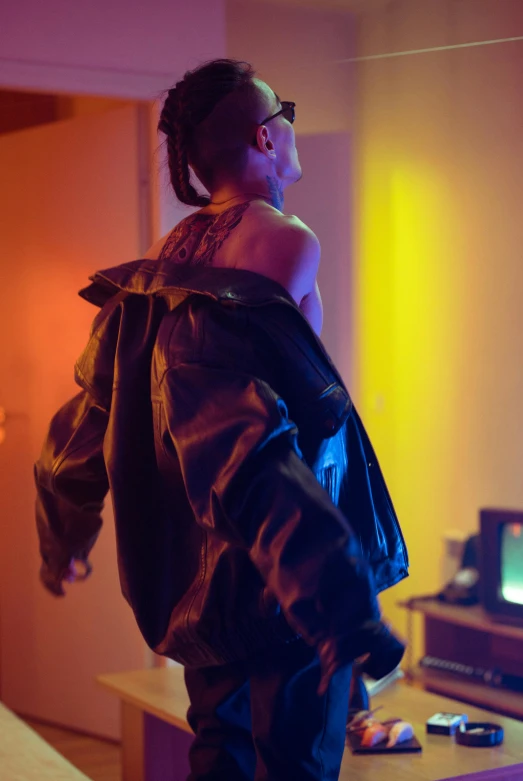 a man standing in a living room next to a tv, inspired by Nan Goldin, trending on pexels, wearing cyberpunk leather jacket, with a ponytail, portrait willow smith, back - lit