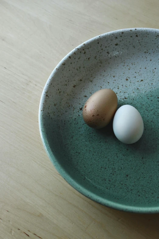 two eggs in a green bowl on a wooden table, a still life, inspired by Maruyama Ōkyo, unsplash, minimalism, on a plate, freckles on chicks, dipped in polished blue ceramic, sustainable materials