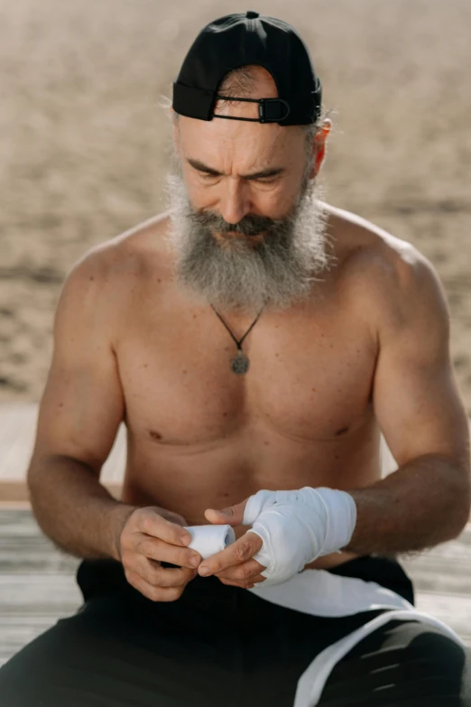 a shirtless man sitting on top of a sandy beach, a tattoo, pexels contest winner, renaissance, white bandage tape on fists, old gigachad with grey beard, holistic medicine advertisement, cardistry