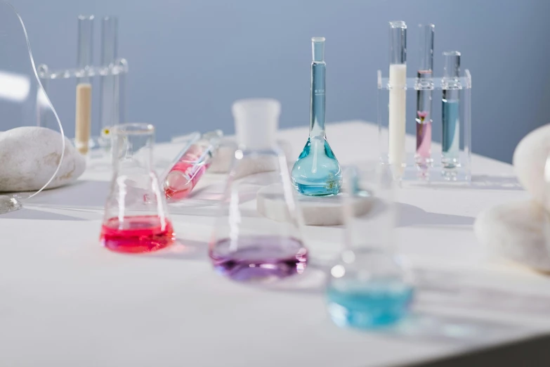 a table topped with lots of different colored liquids, trending on unsplash, clean white lab background, blue and purple colour scheme, flasks, with a lab coat