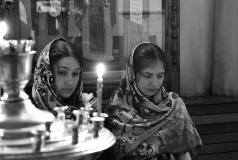 a couple of women sitting next to each other, a black and white photo, by Aleksander Gierymski, flickr, qajar art, candles in foreground, in orthodox church, two girls, 2 0 0 8