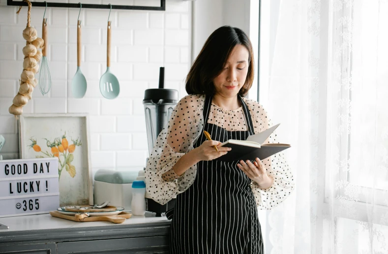 a woman standing in a kitchen reading a book, inspired by Yukimasa Ida, pexels contest winner, best chef, polka dot, malaysian, low quality photo