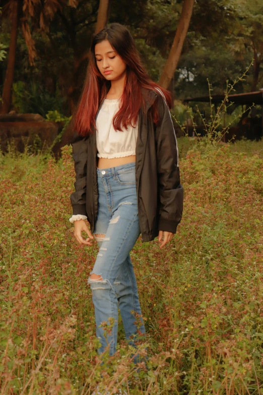 a woman standing in a field of tall grass, an album cover, by Robbie Trevino, trending on pexels, jeans pants, black bomber jacket, long straight red hair, 15081959 21121991 01012000 4k