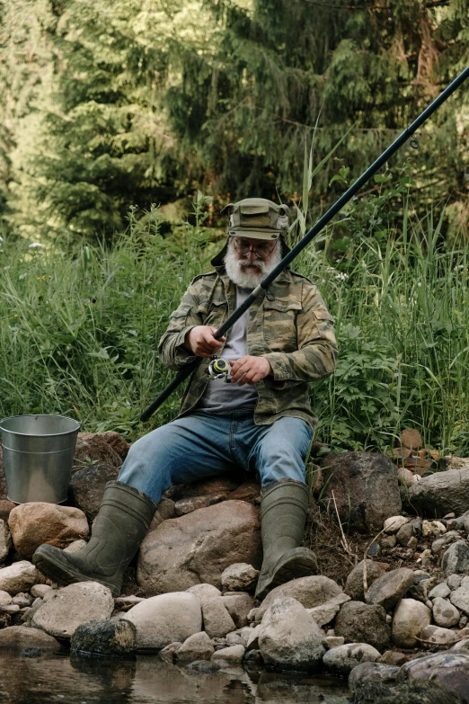 a man sitting on a rock next to a body of water, fishing pole, overalls and a white beard, wearing camo, bill murray