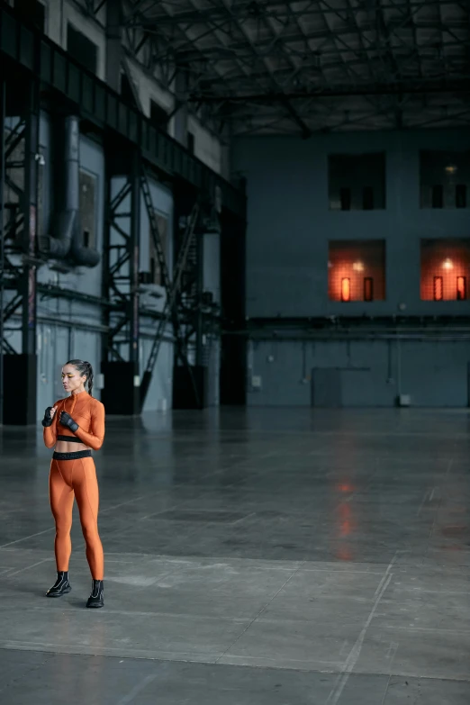 a woman in an orange jumpsuit standing in a building, inspired by Vanessa Beecroft, pexels contest winner, spaceship hangar, wearing fitness gear, solo performance unreal engine, standing in a large empty hall
