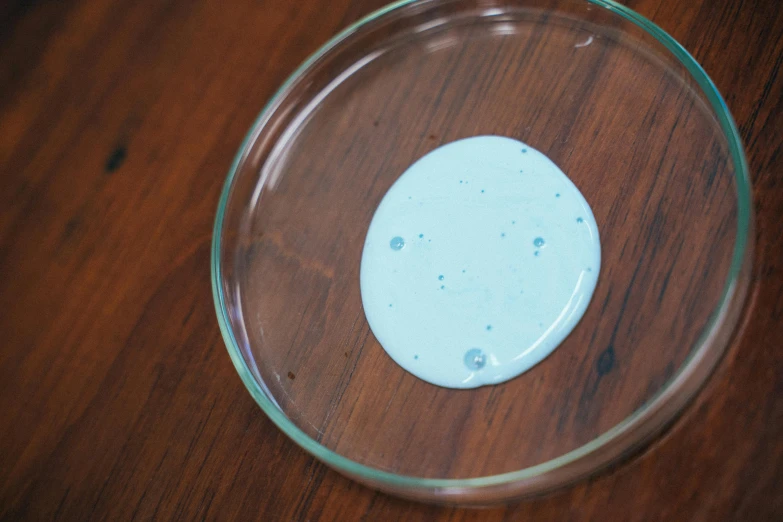 a glass bowl sitting on top of a wooden table, inspired by Damien Hirst, process art, lightblue acrylic paintdrip tar, with a lab coat, birdseye view, escherichia coli