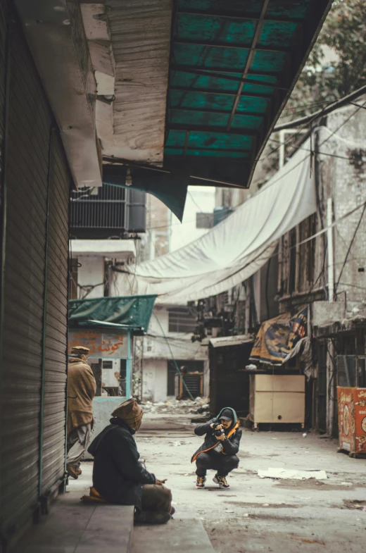 a couple of people sitting on the side of a street, by Ahmed Yacoubi, trending on unsplash, an abandonded courtyard, canopies, collapsed buildings, alley