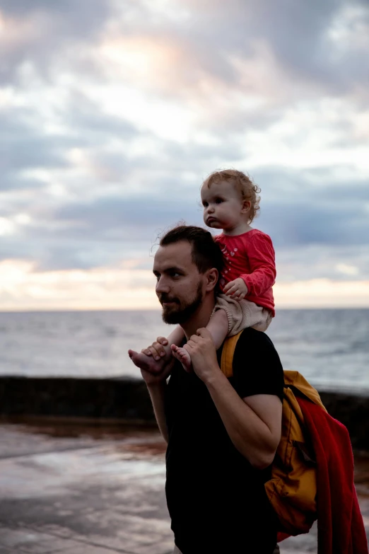 a man carrying a child on his shoulders, by Matt Stewart, unsplash, next to the sea, early evening, bearded, colour photograph