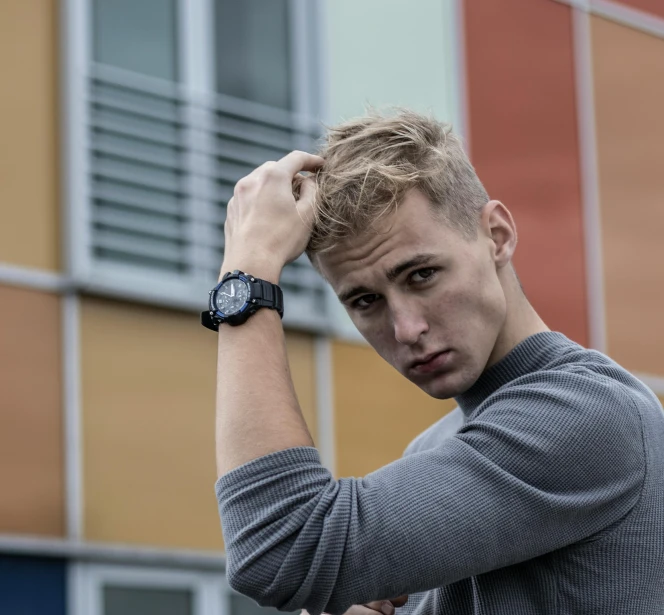 a man standing in front of a building with a watch on his wrist, a portrait, by Andries Stock, pexels contest winner, messy blond hair, non binary model, student, face cluse - up