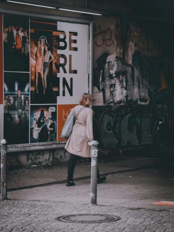 a woman standing on a sidewalk in front of a building, a photo, by Micha Klein, pexels contest winner, happening, there were posters on the wall, berlin city, back of emma stone in beige coat, discovered photo