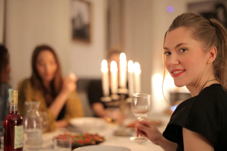 a woman sitting at a table with a glass of wine, candle lighting, people inside eating meals, profile image, lights on