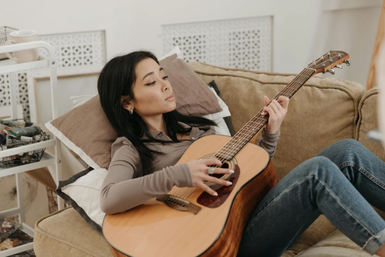 a woman sitting on a couch playing a guitar, inspired by Li Song, trending on pexels, half asian, brown, promotional image, profile pic