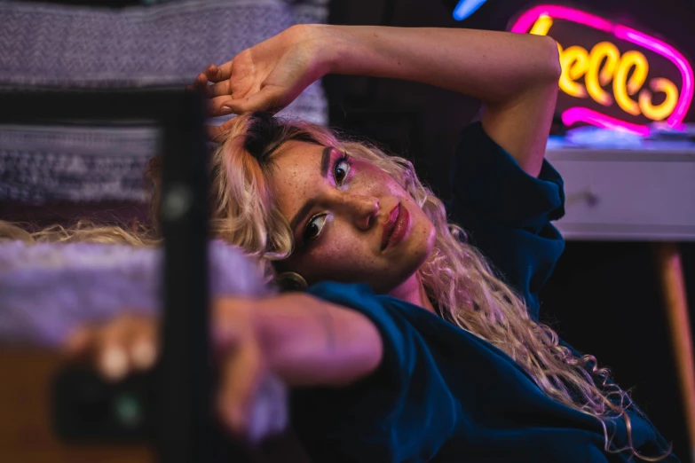 a woman laying on a couch in front of a neon sign, a portrait, inspired by Elsa Bleda, trending on pexels, messy blond hair, sitting at the bar, looking down at you, dua lipa
