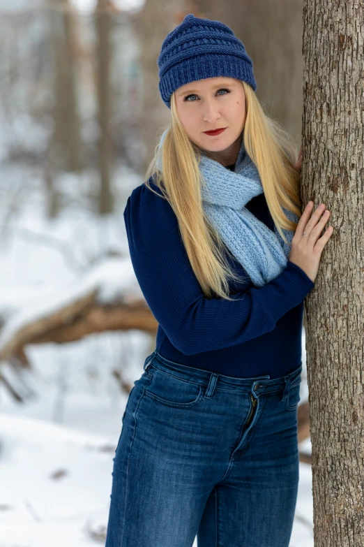a woman leaning against a tree in the snow, a portrait, by Sven Erixson, pexels contest winner, long blonde hair and blue eyes, blue outfit, scarf, blue jeans