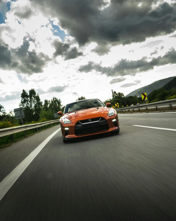 a car driving down a highway under a cloudy sky, by Julia Pishtar, pexels contest winner, nissan gtr r 3 4, orange hue, square, alpine