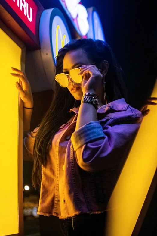 a woman talking on a cell phone next to a neon sign, trending on pexels, yellow carrera glasses, female with long black hair, standing in a dimly lit room, implanted sunglasses