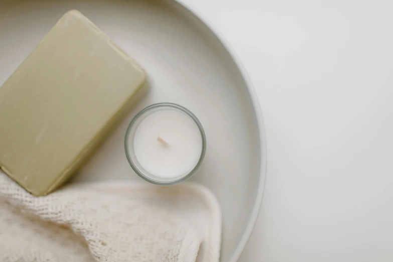 a soap bar sitting on top of a plate next to a candle, trending on pexels, minimalism, sage green, cream white background, high - angle view, thumbnail
