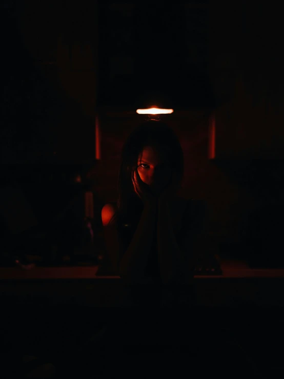 a woman sitting at a table in the dark, red light, wearing a dark hood, in a room, instagram post