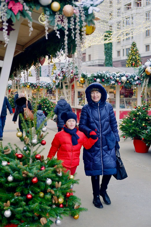 a woman and a child standing in front of a christmas tree, market stalls, walking through the trees, in russia, press shot