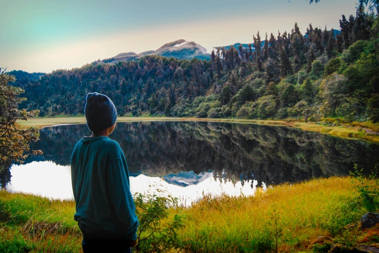 a person standing in front of a lake with a mountain in the background, by Jessie Algie, unsplash contest winner, hurufiyya, haida gwaii, sitting in a colorful forest, profile image, chile