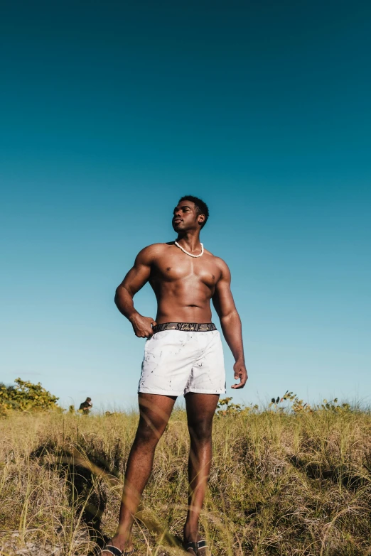 a man standing on top of a grass covered field, bra and shorts streetwear, muscular! white, at a tropical beach, dark skin tone