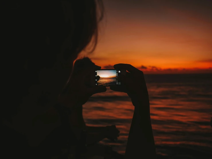 a woman taking a picture of the sunset with her phone, a picture, inspired by Elsa Bleda, pexels contest winner, red and orange glow, on the ocean, holding it out to the camera, night time photograph