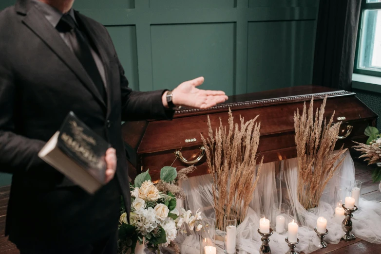 a man in a suit standing next to a casket, trending on unsplash, vanitas, holding spell book, corpse bride style, cottagecore, giving a speech