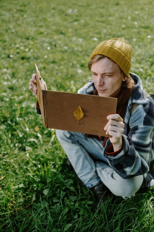 a woman sitting in the grass reading a book, an album cover, inspired by Lasar Segall, trending on pexels, he is wearing a brown sweater, non-binary, caracter with brown hat, he is holding a large book