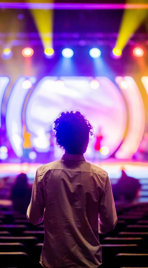 a man standing in front of a stage with bright lights, pexels, happening, east african man with curly hair, people watching around, a round minimalist behind, brightly colored