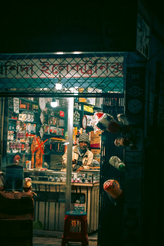 a man sitting in a chair in front of a store, by Jan Tengnagel, unsplash contest winner, bengal school of art, open window at night, at the counter, knick - knacks, vintage color