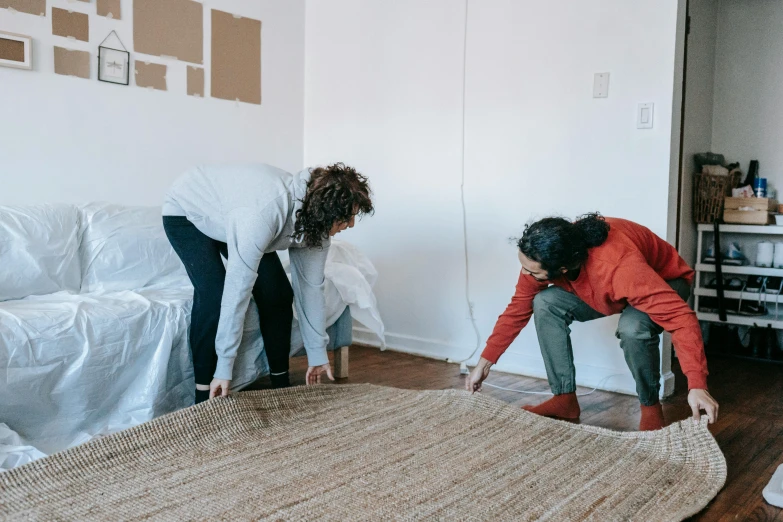 two women working on a rug in a living room, by Carey Morris, pexels contest winner, standing sideways, cardboard, listing image, half - turn