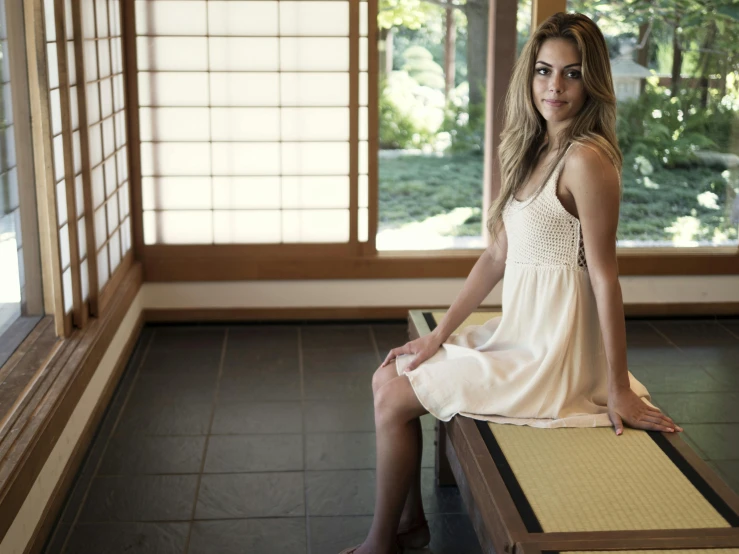 a woman sitting on a bench in a room, a picture, unsplash, mingei, gorgeous female samara weaving, japanese house, wearing ivory colour dress, taken in the late 2010s