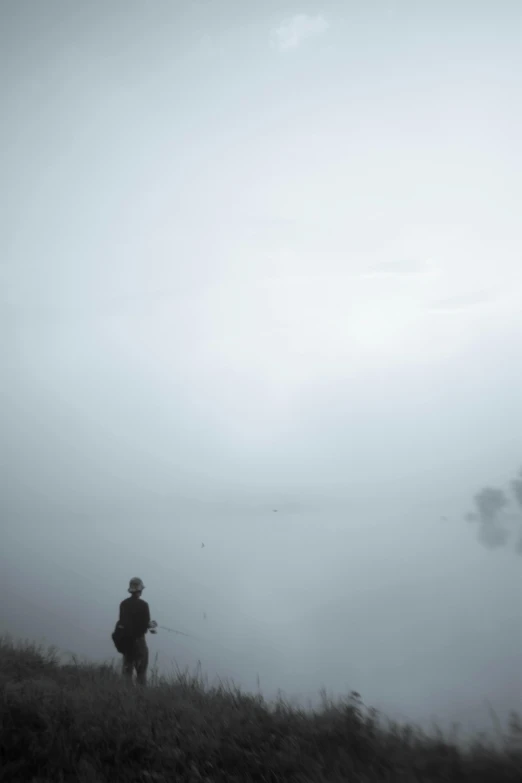 a man standing on top of a grass covered hillside, a picture, by Shang Xi, unsplash, conceptual art, in a foggy pond, fishing, grey, todd hido