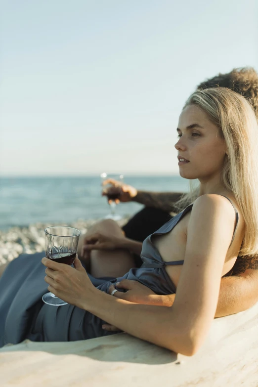 a woman sitting on a beach holding a glass of wine, pexels contest winner, renaissance, couple, young blonde woman, ultrawide cinematic, movie still of a tired