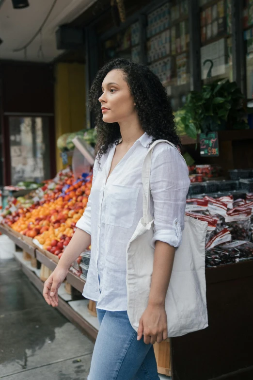 a woman walking down a street past a fruit stand, wearing a linen shirt, thoughtful pose, curly haired, 4l