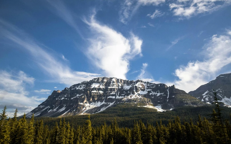 a snow covered mountain with pine trees in the foreground, by Doug Wildey, pexels contest winner, high cliff, cirrus clouds, summer season, slide show