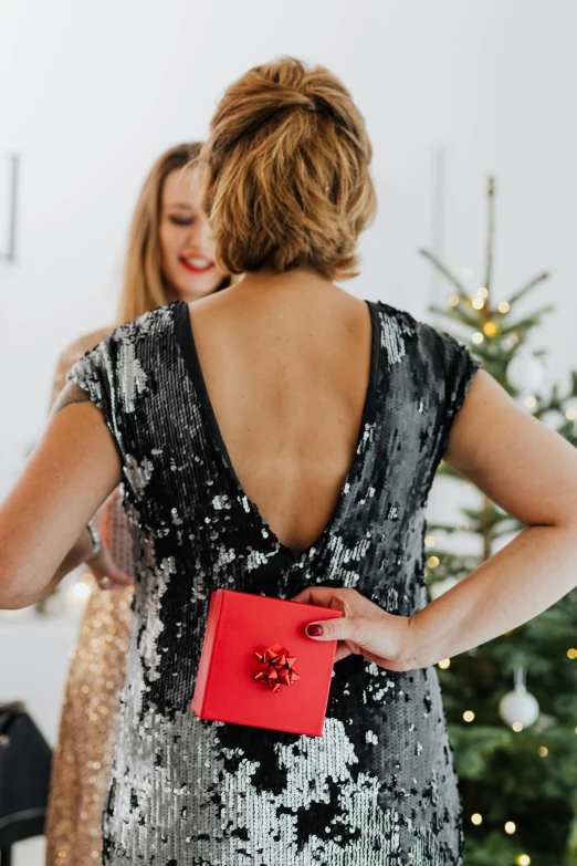 a woman holding a red present in front of a christmas tree, by Julia Pishtar, happening, open back dress, in gunmetal grey, woman holding another woman, wearing black camisole outfit