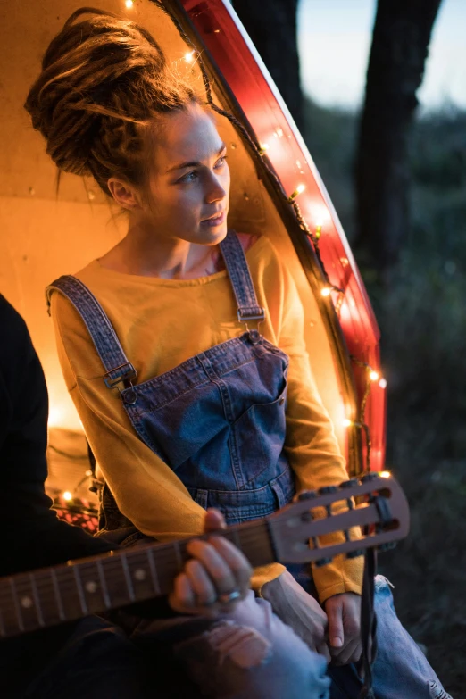 a woman sitting in the back of a van playing a guitar, a portrait, pexels contest winner, happening, wearing overalls, firefly lights, girl with messy bun hairstyle, profile image