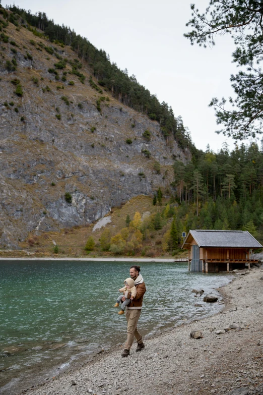 a man standing on a beach next to a body of water, log cabin beneath the alps, father with child, bts, hunting