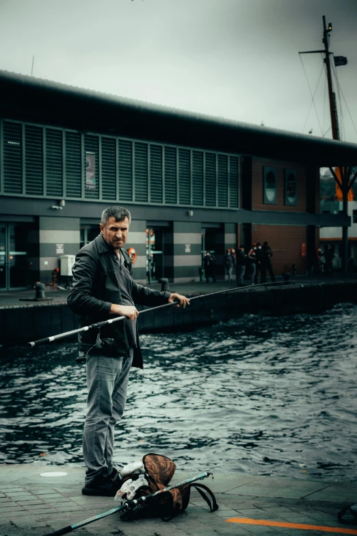 a man standing next to a body of water, fishing pole, in the middle of the city, looking the camera, norwegian man