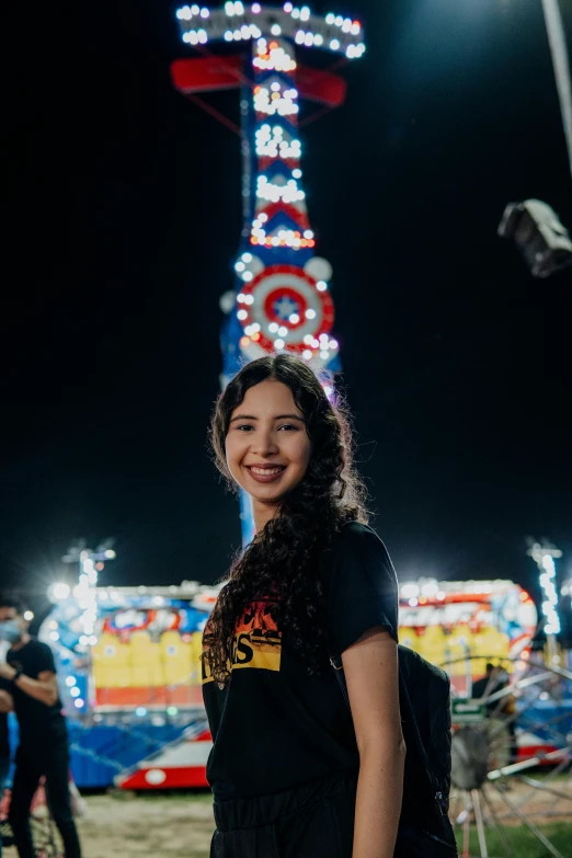 a woman standing in front of a carnival ride at night, by Ric Estrada, pexels contest winner, smiling and looking directly, with towers, karla ortiz, infp young woman