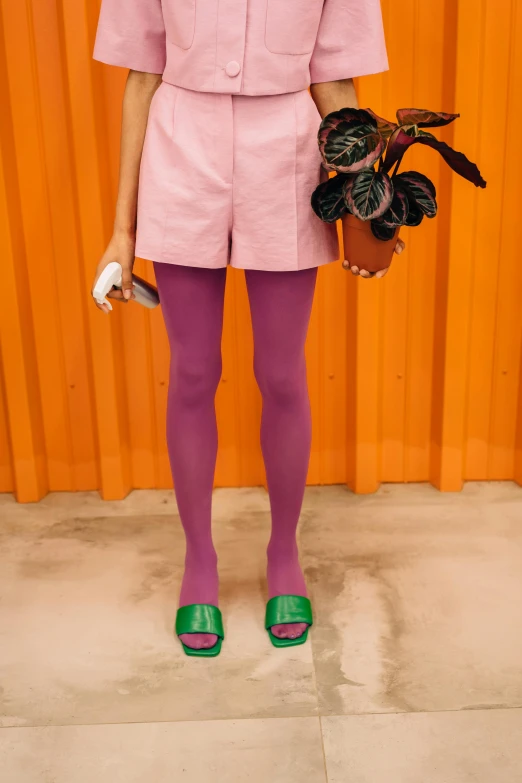 a woman standing in front of an orange wall, an album cover, by Nathalie Rattner, trending on pexels, purple overall shorts, green and pink colour palette, pantyhose tights, ((purple))