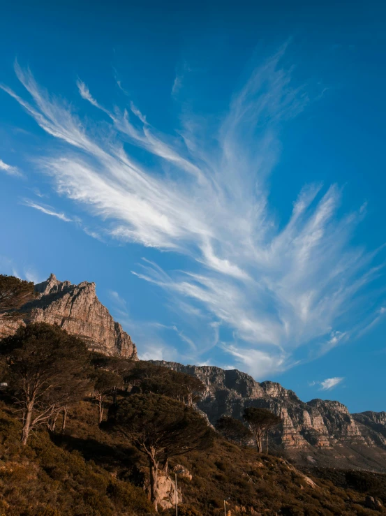 a mountain with some very pretty clouds in the sky, by Arie Smit, wispy clouds in a blue sky, cape, lpoty, trees and cliffs